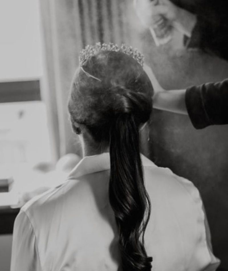 Novia de espalda mientras la están echando la laca en el pelo. Lleva un recogido con coleta y una diadema.